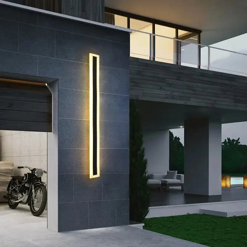 Illuminated vertical black wall light on a gray exterior wall beside a garage